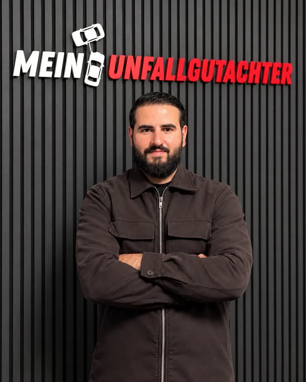 Rojhat Cibakci, Kfz-Sachverständiger bei Mein Unfallgutachter, vor der Logo-Wand im Büro